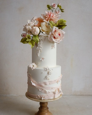 Two-tier white cake with pink and peach gum paste flowers and a fondant ribbon.
