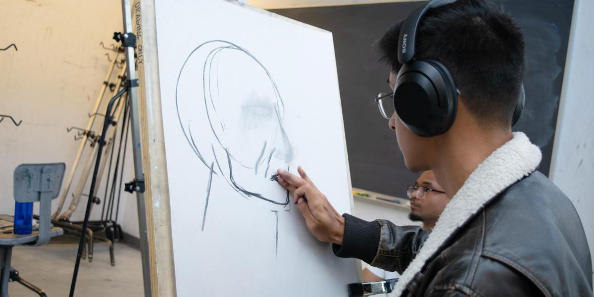 Visual arts student with headphones on drawing a person's face.