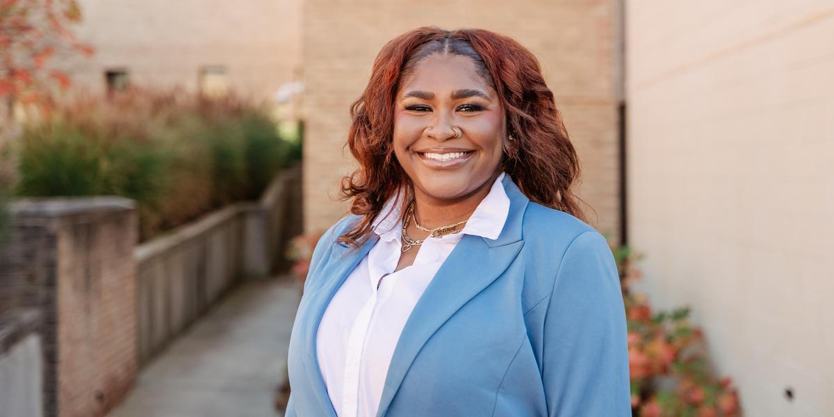 A female student, dressed in a suit, standing on campus and smiling for the camera.