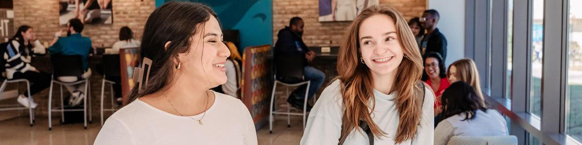 Two students walk and chat while smiling. More students seat at tables behind them.
